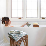 A woman relaxes in a white freestanding bathtub by a large window, holding a folded green floral towel. A matching towel is draped over a woven wooden stool in the foreground, and a wooden bath brush rests on the tub edge.