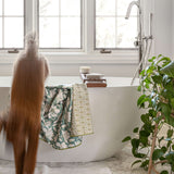 A dog with long light brown hairjumps out of a front of a white freestanding bathtub in a bright bathroom. A floral bath towel and a patterned towel hang from the tub edge, with a wooden tray holding a brush and soap bar.