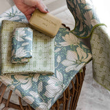 A hand holds a bar of soap over a woven basket holding folded and draped cotton muslin cloths with a floral and geometric pattern in sage green, blue-gray, and cream.