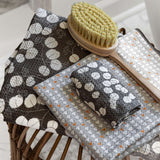 Two patterned fabric washcloths displayed on a wicker stool, with a wooden-handled brush resting on top. One cloth has a dark brown and white circular leaf pattern, and the other has a gray and white geometric pattern with small orange accents.