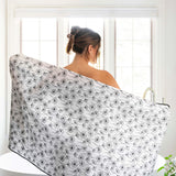 A woman stands with her back to the camera in a bright bathroom, wrapped in a large white muslin towel or wrap printed with a black dandelion pattern. The sink and window are visible in the background.