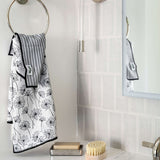 A white hand towel with a black dandelion pattern and a gray-and-white striped folded section hangs from a silver wall ring in a bright bathroom. A mirror, white subway tiles, soap dish, bar of soap, and a wooden-handled brush are visible nearby.