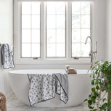 A white freestanding bathtub in a bright bathroom setting, with patterned towels draped over the tub edge and a striped towel hanging on the side. Large white-framed windows, a chrome faucet, and a green houseplant are visible.