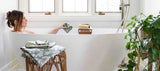 A woman relaxes in a white freestanding bathtub with a floral bath towel draped over the side, set in a bright bathroom with large windows, a plant, and a small wooden stool holding folded towels.