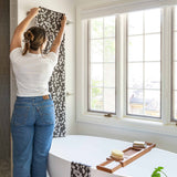 A woman in a white t-shirt and blue jeans hangs a Chocolate brown and white floral patterned bath towel or shower curtain from a rod in a bright, modern bathroom next to a white freestanding bathtub.